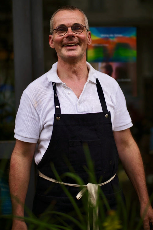 A smiling man wearing glasses, a white short-sleeve polo shirt, and a dark apron standing in front of a softly blurred storefront background.